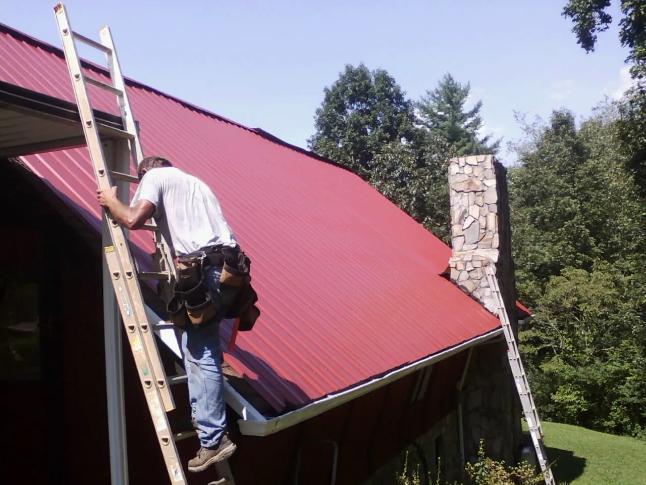 Roofer on a ladder beside a red metal roof and stone chimney — Soffit & Fascia Repair by Roofing Brothers in Coldwater